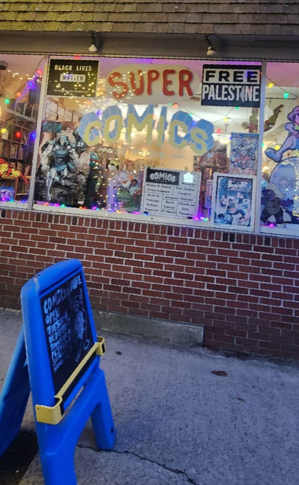The store front window of comics n'more at night. inside the store looks vibnrant and inviting. there's a sign that reads Free Palestine and another that says Super Comics N More. You can see bookcases full of books from the window. 